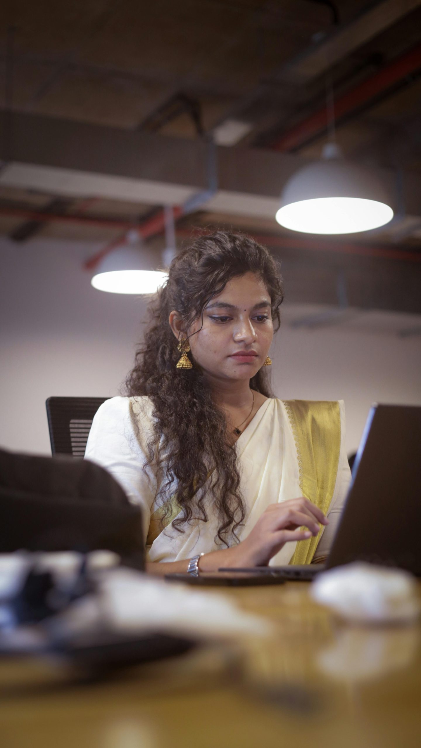 Young South Asian woman in traditional attire working on a laptop in a modern office setting.